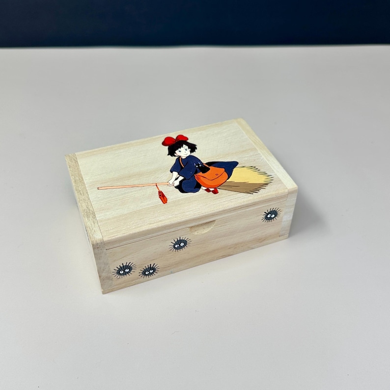 Op de afbeelding: Een lichtgekleurde houten kist met een cartoonillustratie van een jonge heks op een bezem. De kist heeft een scharnierend deksel en is versierd met kleine, zwarte, oogachtige wezens. De heks draagt een blauwe jurk en een oranje tas.