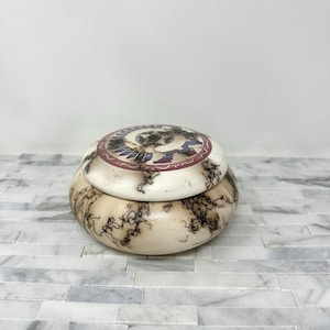 May include: A round, lidded trinket box with a cream-colored base and lid. The lid features a circular design with red, purple, and black accents. The box has a unique, natural pattern of dark lines throughout.