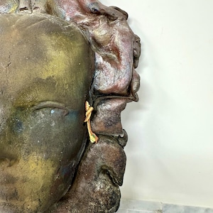 Puede incluir: Primer plano de una escultura texturizada y desgastada de un rostro. El rostro es principalmente marr&oacute;n y dorado, con toques de rojo y azul. Un peque&ntilde;o y ornamentado pendiente dorado cuelga de la oreja. La escultura est&aacute; sobre un fondo blanco.