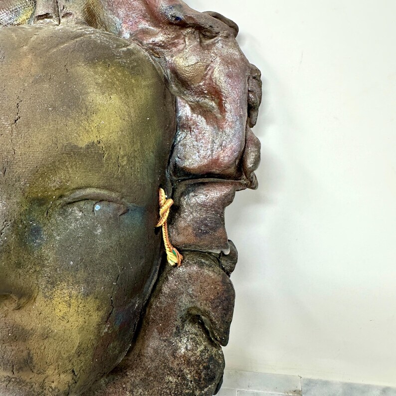 Puede incluir: Primer plano de una escultura texturizada y desgastada de un rostro. El rostro es principalmente marr&oacute;n y dorado, con toques de rojo y azul. Un peque&ntilde;o y ornamentado pendiente dorado cuelga de la oreja. La escultura est&aacute; sobre un fondo blanco.