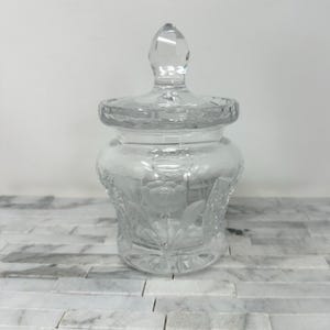 May include: A clear glass jar with a lid. The jar features an etched floral design and a faceted knob on the lid. The jar is sitting on a white and gray surface.