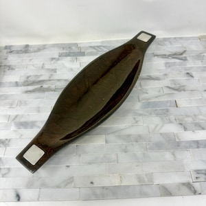 May include: A dark brown, elongated decorative bowl with a glossy finish. The bowl has a boat-like shape with a rectangular silver accent at each end. The interior of the bowl is a darker shade of brown.