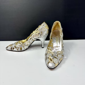 May include: A pair of silver and gold high-heeled shoes with intricate cut-out designs. The shoes feature a pointed toe and a stiletto heel. Embellishments include small, sparkling details. The shoes are from the brand Helene Arpels.