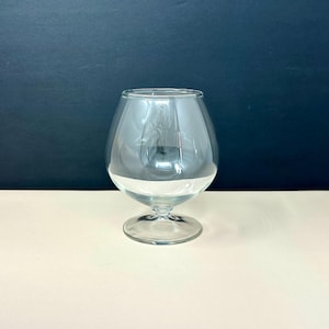 May include: A clear, stemmed brandy snifter glass. The glass has a rounded bowl and a short stem with a circular base. The glass is transparent, allowing light to pass through. The background is a dark blue and cream color.