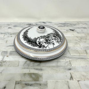 May include: A white ceramic lid with a black and gold design. The lid features two figures in black and white, with a gold trim around the edge. The lid has a knob on top.