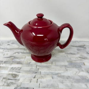 May include: A deep red ceramic teapot with a lid and a handle. The teapot has a rounded body and a small base.