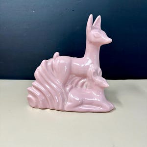 May include: A glossy, light pink ceramic figurine depicting two deer. One deer stands tall with ears pointed upward, while the second smaller deer rests in a lying position directly in front of the standing deer. The piece has a smooth finish.