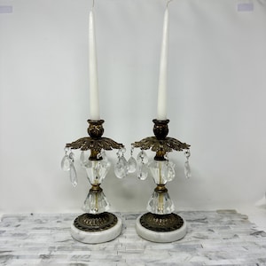 May include: Two ornate candle holders with white candles. The holders have a gold-colored metal base and accents, with clear crystal embellishments and hanging teardrop-shaped crystals. The base is white marble.