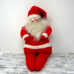 May include: A plush Santa Claus doll with a white beard and red and white suit. The doll is sitting with its legs crossed.