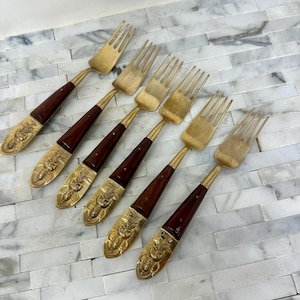 May include: Set of six gold-tone forks with brown wooden handles. The forks have intricate designs on the handles.