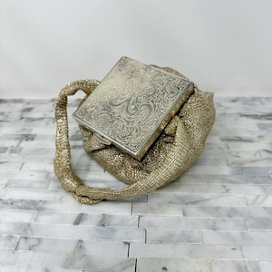 May include: A vintage silver compact with an ornate floral design rests on a gold mesh handbag. The compact is rectangular and has a clasp. The handbag has a twisted handle and a gathered body. The background is a marble surface.