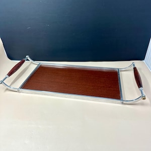 May include: A rectangular serving tray featuring a dark brown wood-grain surface framed by a silver-toned metal border. The tray includes two curved silver metal handles, each accented with a smooth, dark brown wooden grip on either side.