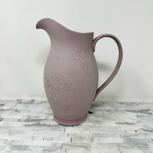 May include: A dusty rose-colored pitcher with a curved handle and spout. The ceramic pitcher features etched floral designs on the front and top. The pitcher is a decorative item.