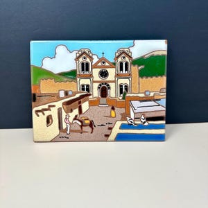 May include: A square ceramic tile featuring a colorful illustration of a church and surrounding buildings. The artwork depicts a sunny day with a blue sky, clouds, and green hills in the background. A person and a donkey are in the foreground.