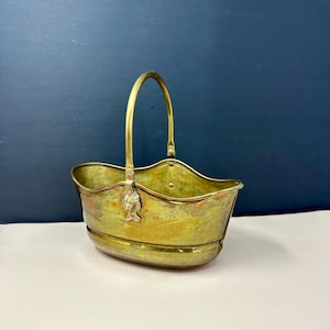 May include: A vintage brass basket with a curved handle and decorative fish-shaped accents. The basket has a rounded base and a wavy rim. The brass has a warm, aged patina, suggesting its antique nature. The basket is likely used for decorative purposes.