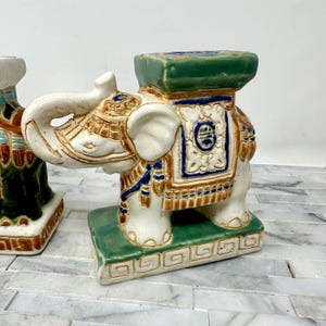 May include: A ceramic elephant garden stool with a white body, brown and blue accents, and a green seat. The elephant has detailed carvings and a decorative saddle. The stool is placed on a green base with a geometric pattern.
