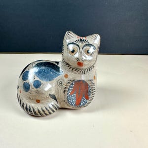May include: A ceramic cat figurine featuring a cream-colored base with hand-painted details. The cat has blue leaf-like patterns on its back, a red floral design on its chest, and black striped markings on its tail. It has large eyes and a red nose.