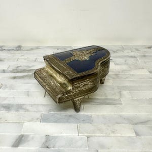 May include: A vintage piano-shaped trinket box. The lid is navy blue with a gold-colored embossed design. The body and legs are gold-toned metal. The box is a decorative item.