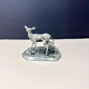 May include: A silver-colored figurine of a doe and her fawn standing on a textured base. The doe stands tall, while the fawn is smaller with spotted markings. The base has a naturalistic appearance, suggesting a forest setting. The figurine is a detailed representation of wildlife.