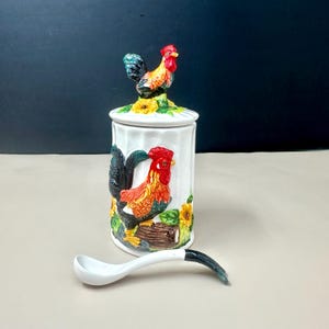 May include: A white ceramic jar with a rooster design, featuring a red comb and colorful feathers, with a matching spoon. The jar has a lid with a rooster figurine on top and is decorated with sunflowers.