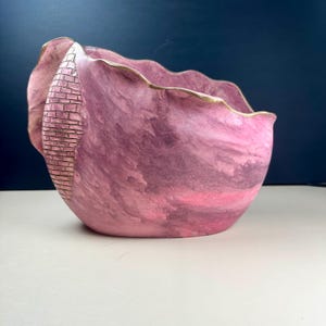 May include: A bowl with a wavy, scalloped rim and a marbled pink and purple finish. One side features a vertical, textured panel with a raised brick-like pattern. The top edge of the bowl is accented with a thin, metallic gold-coloured trim.