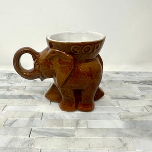 May include: Brown ceramic elephant-shaped mug with a white interior. The mug has a handle shaped like an elephant's trunk and the letters "GOP" on the rim. A novelty item.
