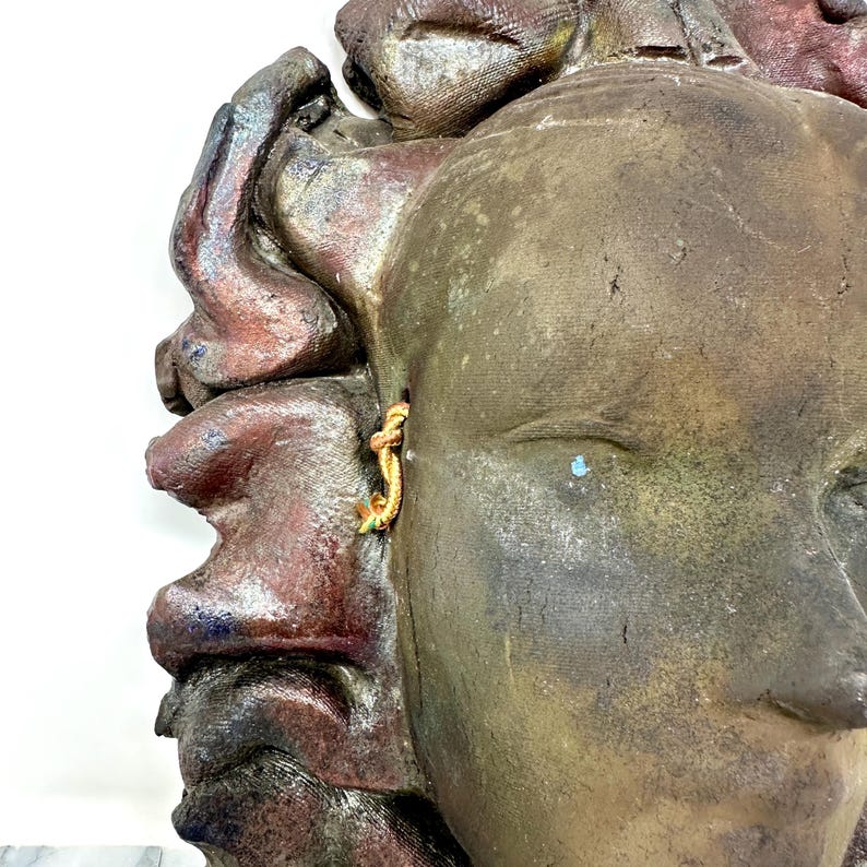 Puede incluir: Primer plano de una escultura de cer&aacute;mica que representa un rostro con una superficie texturizada. La escultura presenta un pendiente dorado y es principalmente marr&oacute;n con toques de rojo y azul.