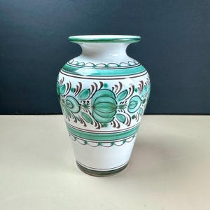 May include: A white ceramic vase featuring a wide, flared rim and a tapered base. The exterior is decorated with horizontal bands of green and brown, including a central pattern of stylized green onion-like motifs and brown leaf accents.
