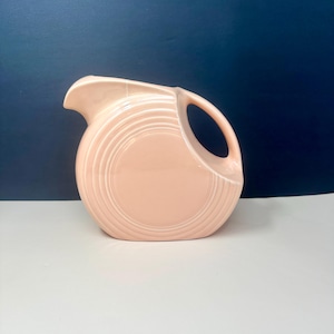 May include: A vintage, blush-colored pitcher with a circular design. The pitcher has a curved spout and a large, open handle. The body of the pitcher features concentric, ridged detailing. The background is a dark blue.