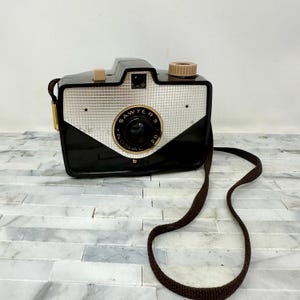May include: Vintage Sawyer's Nomad 20 camera. The camera has a black body with a silver and black front panel. The lens is surrounded by the text "Sawyer's Nomad 20". It has a brown strap and tan-colored buttons.