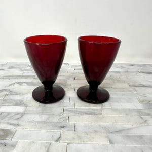 May include: Two ruby red glass goblets with a conical bowl and a circular base. The stem and base are a darker shade of red. The glasses are displayed on a white and gray tiled surface.