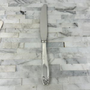 May include: A silver butter knife with a floral design at the base of the handle. The knife has a long, flat blade and a rounded handle. The handle has a decorative floral pattern.