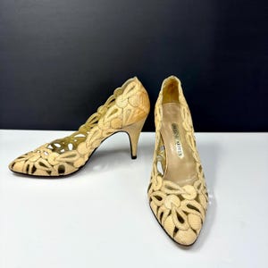 May include: A pair of beige high-heeled shoes with intricate cut-out floral designs. The shoes have pointed toes and slender heels. The brand name "Helene Arpels" is visible on the insole. The shoes are made of a textured material.