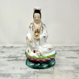 May include: A seated porcelain figurine of a woman in a white robe with gold accents, holding a golden vase. The figure is atop a lotus flower base with green, pink, and white colors. The figurine has a serene expression.