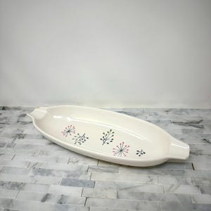 May include: Cream-colored, oblong ceramic serving dish with a floral pattern in red, black, and green. The dish has a handle on each end. A decorative piece for serving food.