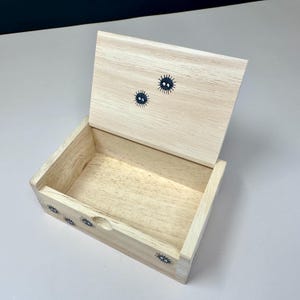 Op de afbeelding: Een lichtgekleurde houten kist met een open deksel. De kist is versierd met kleine, zwarte, ronde karakters met stekelige details. De kist is leeg en staat op een wit oppervlak.