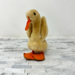 May include: A vintage plush duck toy with a tan body, orange beak, and orange feet. The duck has a fluffy texture and small black eyes. The toy is standing on a white and gray marble surface.