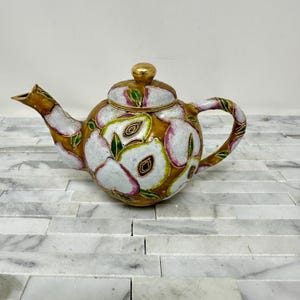 May include: A white ceramic teapot with a gold lid and a floral design. The teapot is decorated with pink and green flowers and has a gold trim.