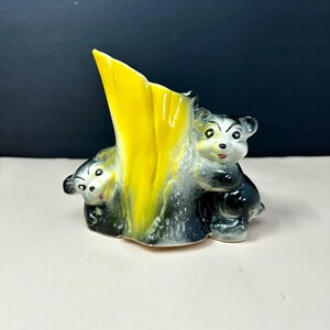 May include: A ceramic vase featuring a bright yellow central section shaped like a tree trunk, flanked by two small black and white bear figures. The bears have painted facial features, and the base of the vase is textured to resemble bark.