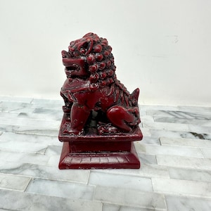 May include: A red Foo Dog statue, a traditional Chinese guardian figure, sits on a square base. The statue has a detailed mane, a fierce expression, and is in a seated position. The statue is made of a solid material and has a glossy finish.