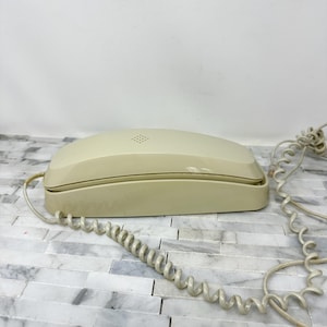 May include: A vintage cream-colored telephone with a coiled cord. The phone has a rectangular base with a rounded top and a small speaker grille. The coiled cord is connected to the base and extends to the right.