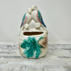 May include: A vintage ceramic wall pocket featuring two perched birds with white, blue, and pink accents. The cream-colored pocket has a leaf design in teal and brown. This decorative piece is ideal for adding a touch of nature-inspired charm to any space.