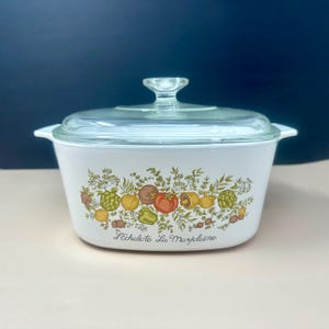May include: White casserole dish with a clear glass lid and handle. The dish features a colorful floral design with vegetables and the text "L'échalote La Marjolaine". A vintage kitchenware item.