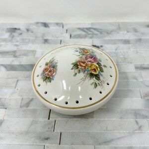May include: A white ceramic pincushion with a floral design featuring pink, yellow, and blue roses. The pincushion has a gold rim and a domed top with holes for pins.