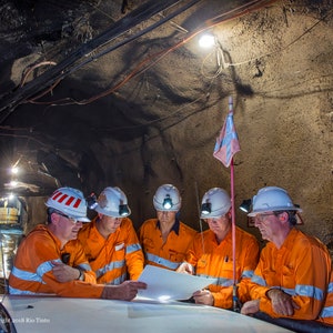 Może przedstawiać: Pięciu mężczyzn w kaskach i pomarańczowych kamizelkach bezpieczeństwa stoi w ciemnym tunelu i patrzy na kartkę papieru. Tunel jest oświetlony światłami sufitowymi, a z sufitu zwisa flaga.