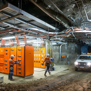 Może przedstawiać: Widok podziemnej kopalni z pomarańczowymi panelami elektrycznymi, białym pickupem i dwoma pracownikami w kaskach i kamizelkach bezpieczeństwa.