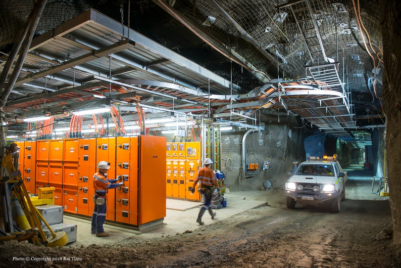 Może przedstawiać: Widok podziemnej kopalni z pomarańczowymi panelami elektrycznymi, białym pickupem i dwoma pracownikami w kaskach i kamizelkach bezpieczeństwa.