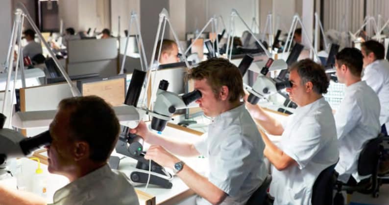 Może przedstawiać: Grupa os&oacute;b pracujących przy indywidualnych stanowiskach pracy z mikroskopami. Każda osoba ma na sobie biały fartuch laboratoryjny i skupia się na swojej pracy.