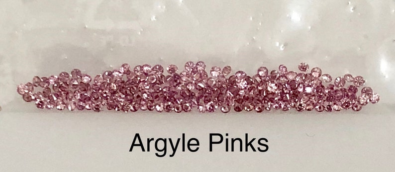 Może przedstawiać: Zbliżenie na kolekcję małych, r&oacute;żowych diament&oacute;w Argyle. Diamenty są luźne i rozrzucone na białym tle. Pod diamentami widnieje napis "Argyle Pinks".