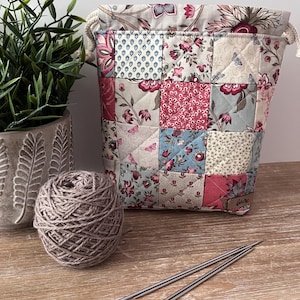 May include: A quilted drawstring pouch featuring a patchwork design of floral and geometric patterns in shades of pink, blue, and cream. The bag has white rope drawstrings and a small brown rectangular label on the lower right corner.